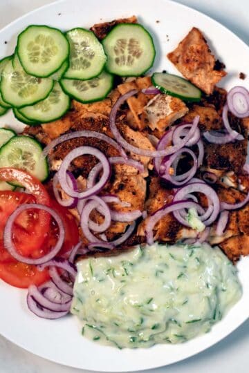 A big serving of Shaved Tofu Gyros on a plate with tzatziki, onions, tomatoes, and cucumber slices.