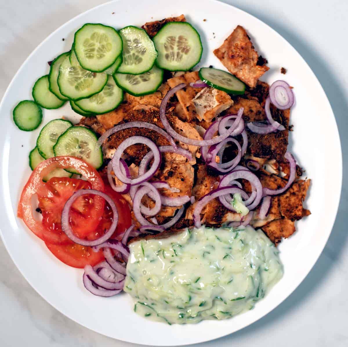 A big serving of Shaved Tofu Gyros on a plate with tzatziki, onions, tomatoes, and cucumber slices.
